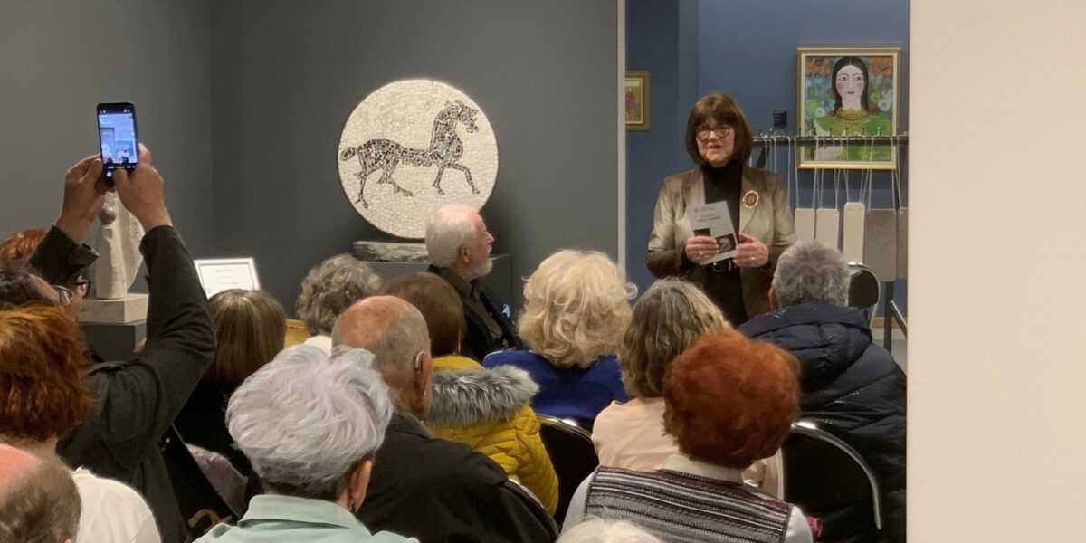 Odprtje razstave Odtisi v kamnu v Galeriji Trebnje. Veliko ljudi sedi na stolih, umetnica Jožica Škof stoji in nagovarja ljudi.