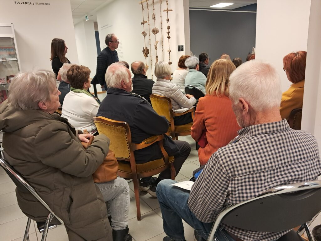Odprtje razstave Odtisi v kamnu. Ljudje sedijo in poslušajo glasbeni nastop.