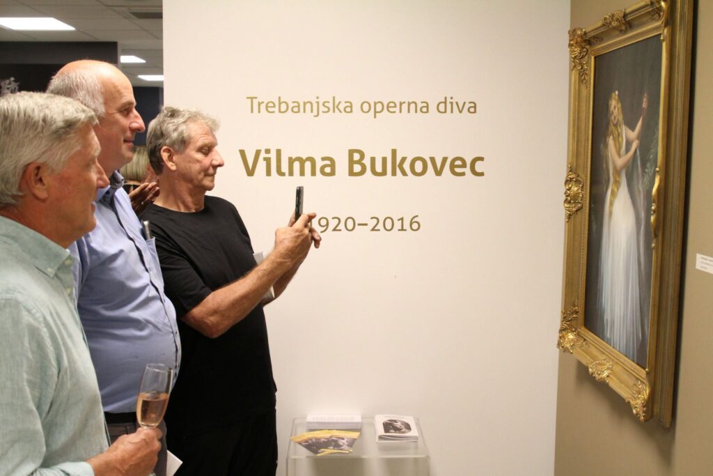 Trije moški si na otvoritvi ogledujejo sliko in jo fotografirajo z mobitelom.