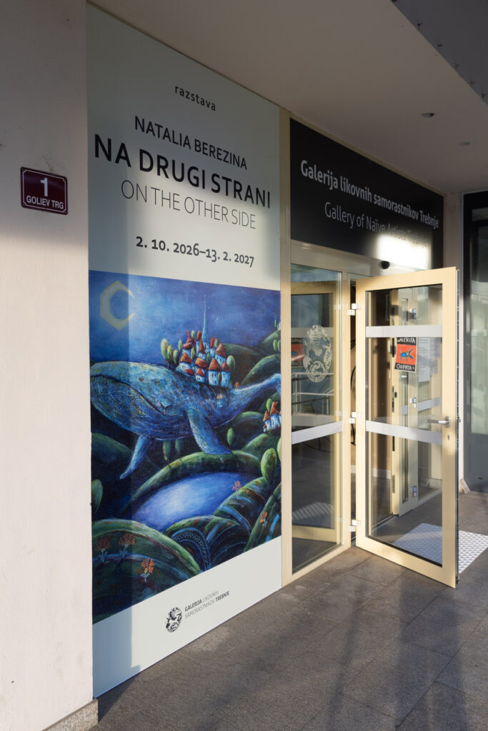 Vrata v Galerijo likovnih samorastnikov Trebnje so odprta, nad njimi je ime galerije v slovenščini in v angleščini. Levo je velik reklamni pano za razstavo Natalia Berezina Na drugi strani. Spodaj je pripisan še angleški naslov On the Other Side. Opazna je nosilna podoba z kitom, ki nosi več hiš na svojem hrbtu in plava nad hribi in dolinami. Spodaj je logotip galerije. Vidna je hišna številka Goliev trg 1.